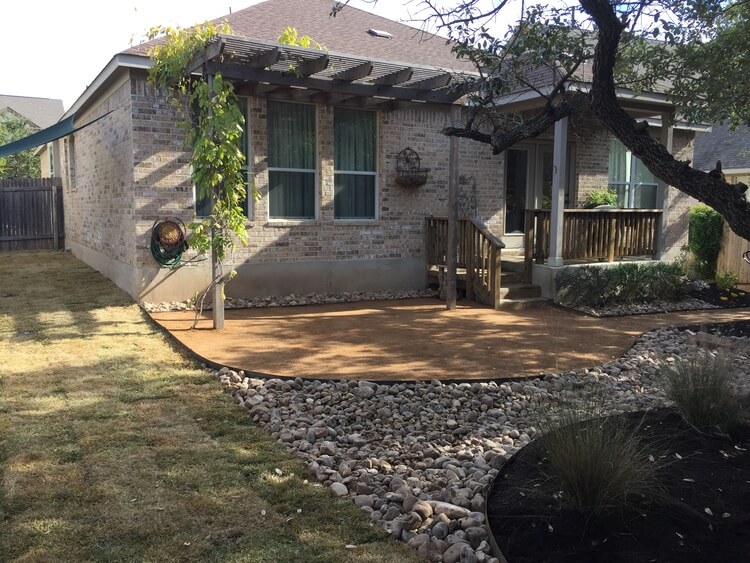 backyard with new grass, a section of gravel, a section of pebbles, and a dirt plant bed. A house with a wooden deck can be seen in the background.